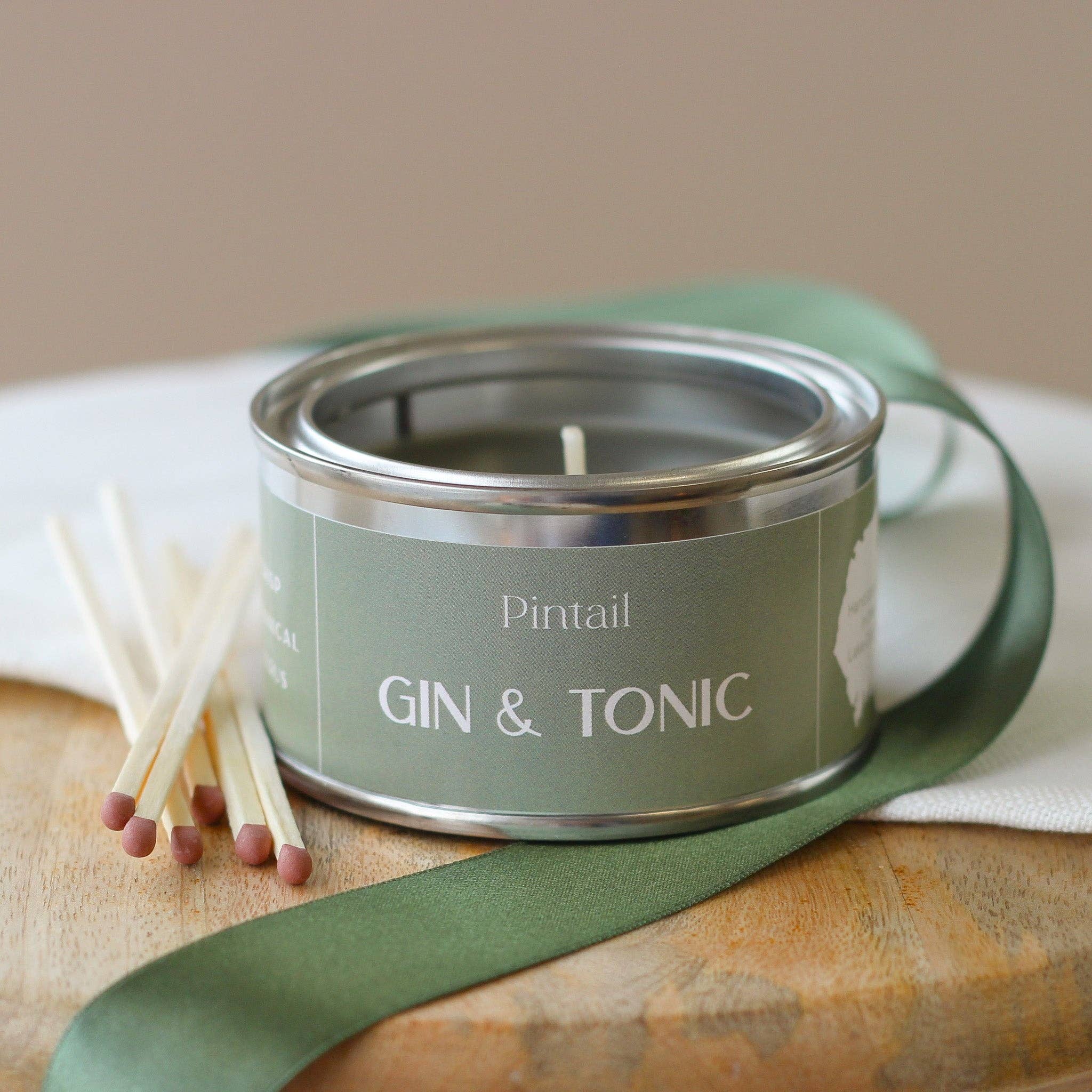 Candle in a tin labeled 'GIN & TONIC' with matches on a wooden surface.