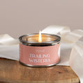 Candle in a tin labeled 'Trailing Wisteria' by Pintail on a wooden surface with white fabric.