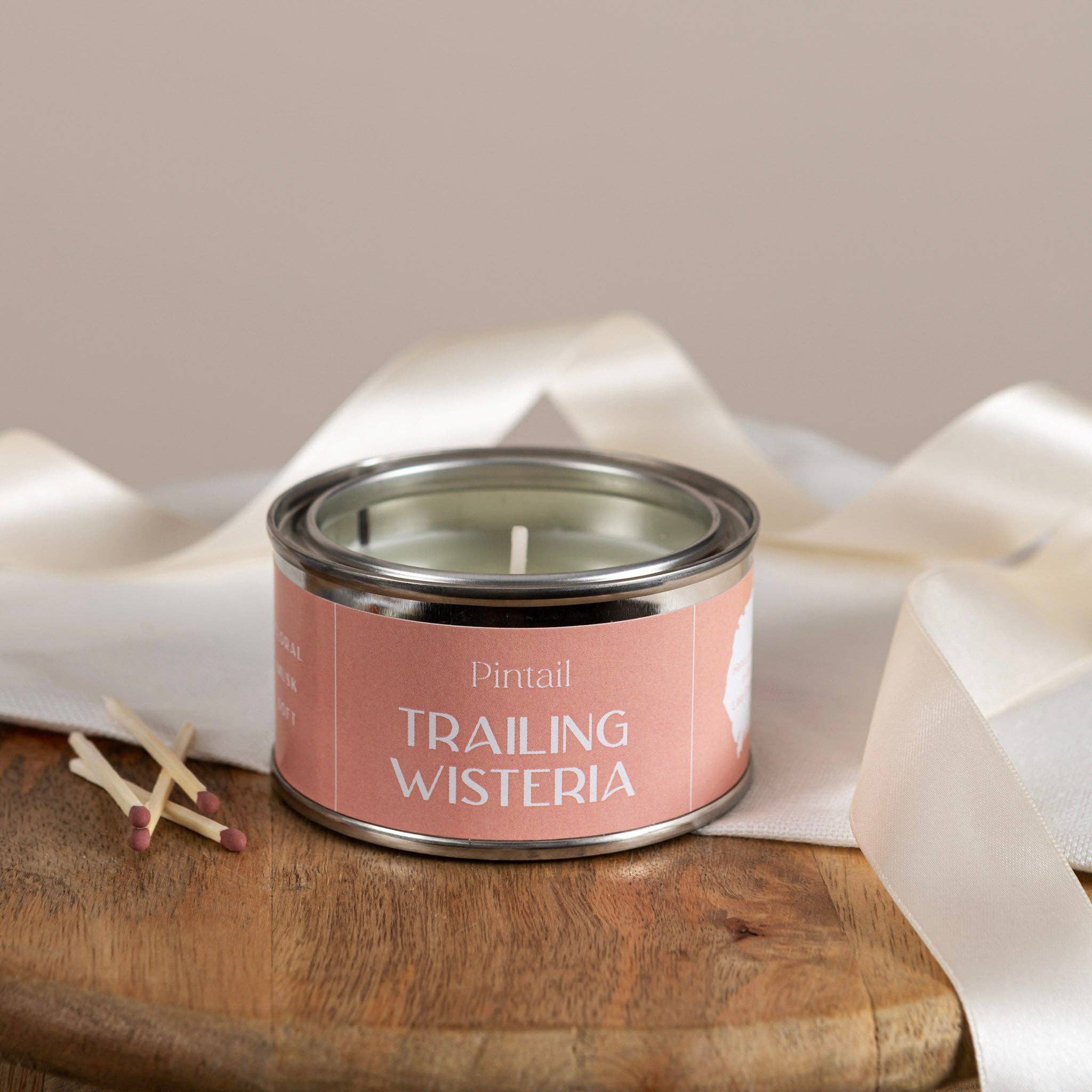 Candle in a metal tin labeled 'Trailing Wisteria' on a wooden surface with white fabric.